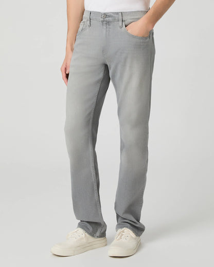 A person wears Paige Grey Wainwright Normandie Regular Fit Jeans with a white shirt and white sneakers. One hand is in their pocket. The visible lower torso and legs are set against a plain light gray background. , available at Richard Gelding Mayfair