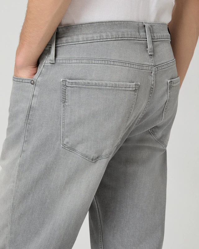A close-up of a person wearing Paige Grey Wainwright Normandie Regular Fit Jeans, shown from the waist down and from behind with one hand in the back pocket. The jeans feature visible stitching, belt loops, and are paired with a white shirt against a plain light gray background. , available at Richard Gelding Mayfair