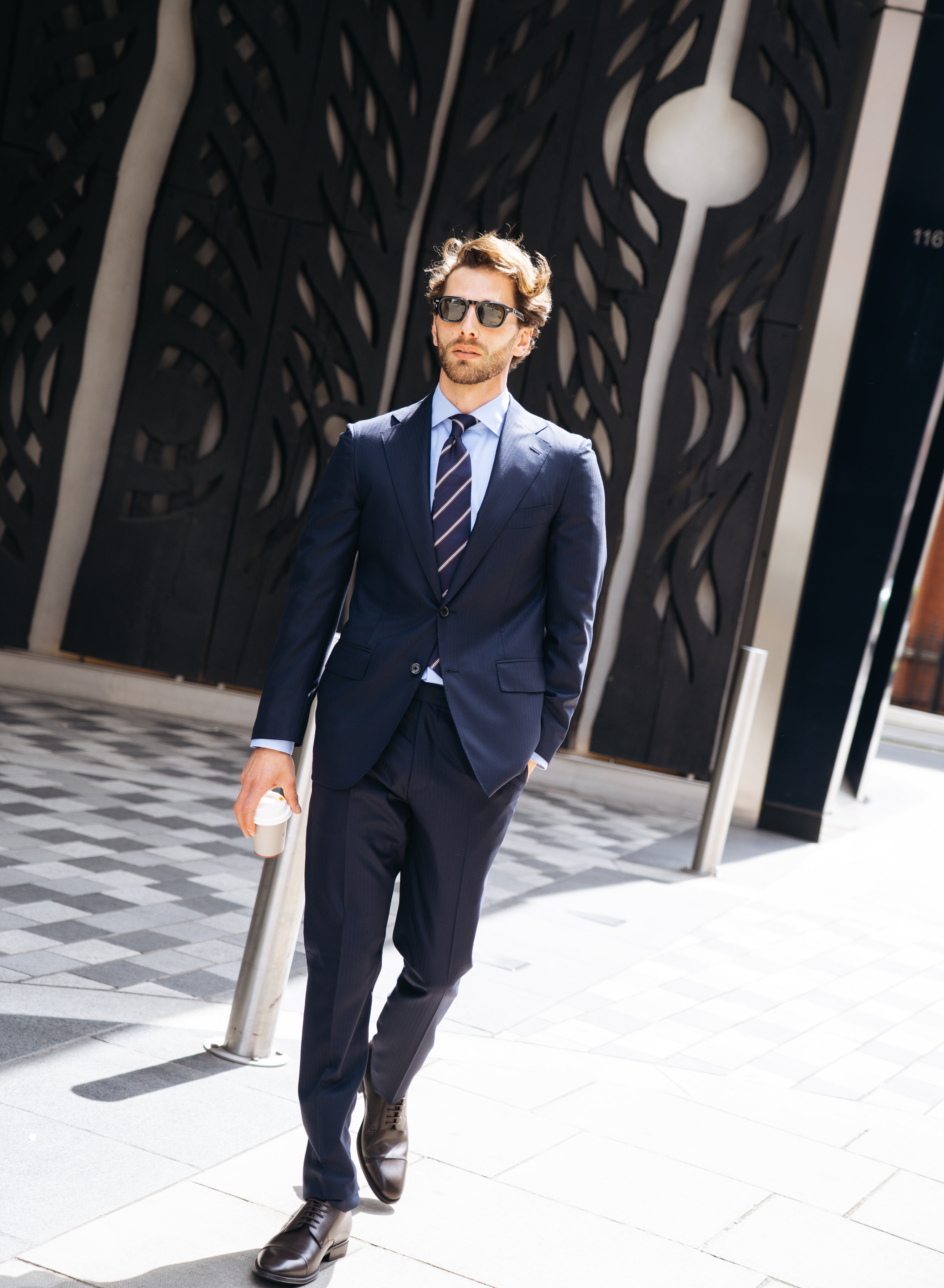 A man in a Cavour Navy Herringbone Super 150's Wool Suit walks confidently outside on a sunny day, wearing sunglasses, dress shoes, and carrying a coffee cup. A modern black wall with abstract patterns stands behind him. , available at Richard Gelding Mayfair