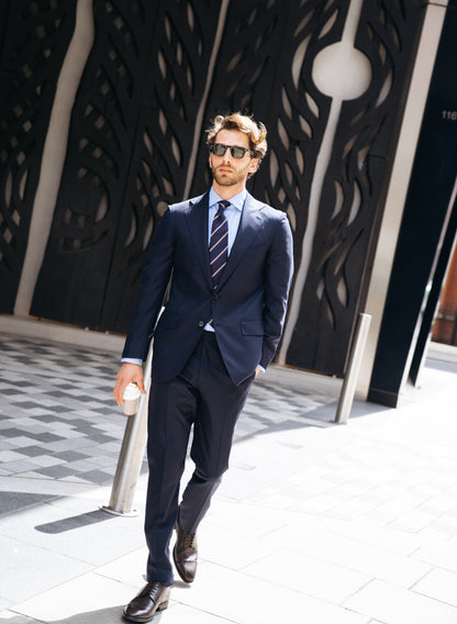 A man in a Cavour Navy Herringbone Super 150's Wool Suit walks confidently outside on a sunny day, wearing sunglasses, dress shoes, and carrying a coffee cup. A modern black wall with abstract patterns stands behind him. , available at Richard Gelding Mayfair