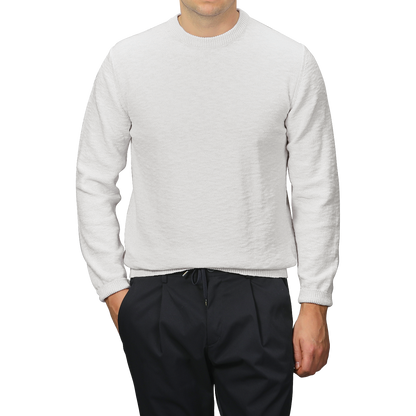A person wearing a Maurizio Baldassari Cream Summer Mouline Crew Neck Sweater and black pants stands against a gray background, with hands partially in their pockets, highlighting the comfortable cotton-viscose blend.