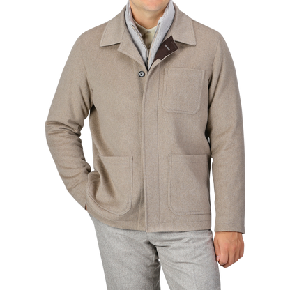 A person is wearing a Beige Cashmere Half Padded Chore Coat by Maurizio Baldassari with three pockets over a light grey shirt, complemented by grey trousers. The ensemble features a detachable wool-cashmere waistcoat peeking out, adding an extra layer of sophistication. Their head is not visible in the image.