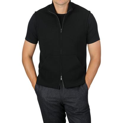 Here’s a revised version of the sentence incorporating the given product data:

A person wearing a black short-sleeve shirt under the Maurizio Baldassari Black Milano Stitch Wool Zip Gilet pairs it with dark gray pants, with hands partially in the pants pockets. The background is plain light gray.