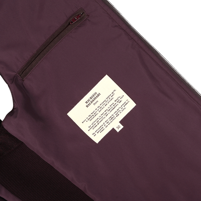 A close-up of the Maurizio Baldassari Plum Water Repellent Cashmere Gilet reveals its rich, dark purple hue. The interior label displaying care instructions is prominently featured, along with a small zippered pocket next to it.