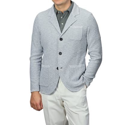 A man exudes relaxed luxury in the Sky Blue Summer Mouline Knitted Jacket by Maurizio Baldassari, paired with a dark gray collared shirt and white pants, one hand casually in his pocket. The cotton-viscose blend enhances sophistication against the plain background.