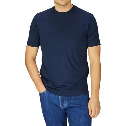 Man wearing a Mazzarelli navy blue merino wool T-shirt and blue jeans, standing with one hand slightly tucked in his pocket, against a striped grey background.