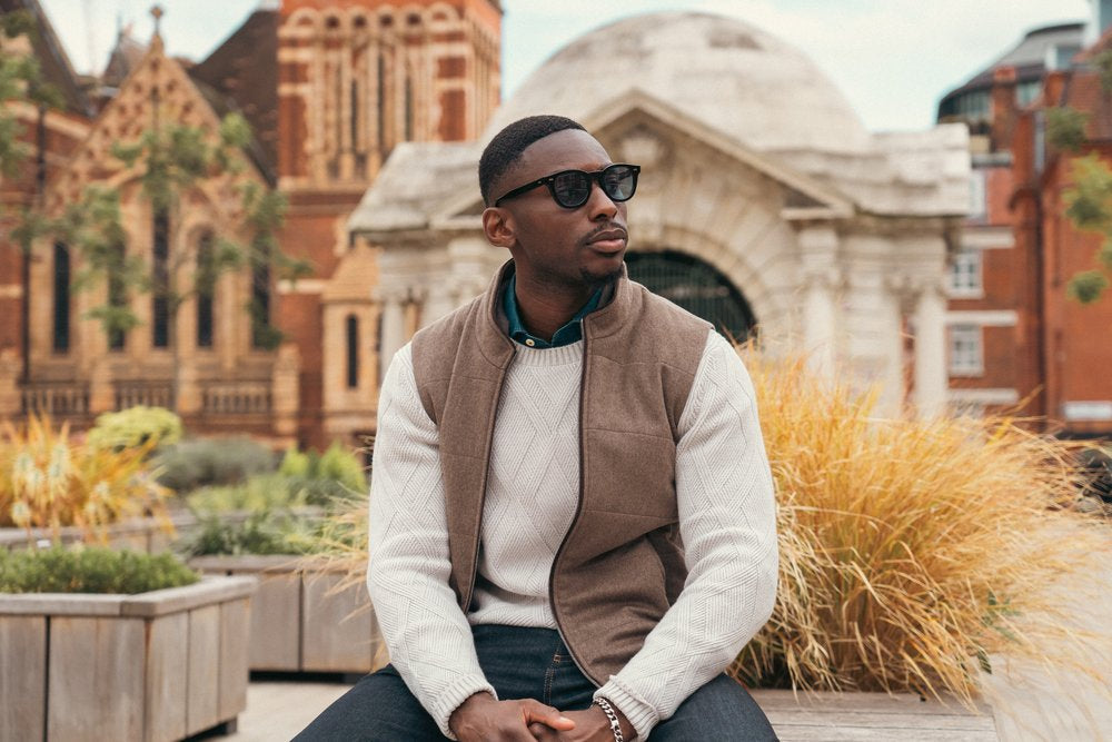 A stylish man in sunglasses, a tan vest, and a cream sweater sits outdoors on a bench. Behind him are historic brick buildings, an arched stone structure, and ornamental grasses, creating an urban, autumn atmosphere. , available at Richard Gelding Mayfair