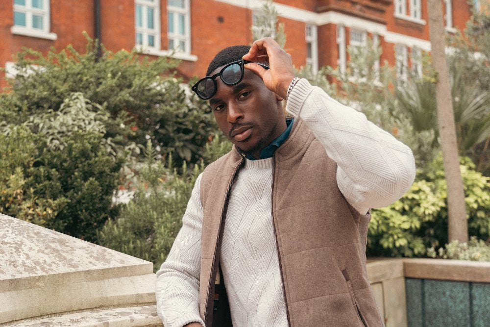 A man outdoors adjusts his sunglasses while looking at the camera. He wears a white sweater under a light brown vest. Behind him are green bushes, trees, and a red brick building with white window frames. The setting appears to be in a city or campus. , available at Richard Gelding Mayfair