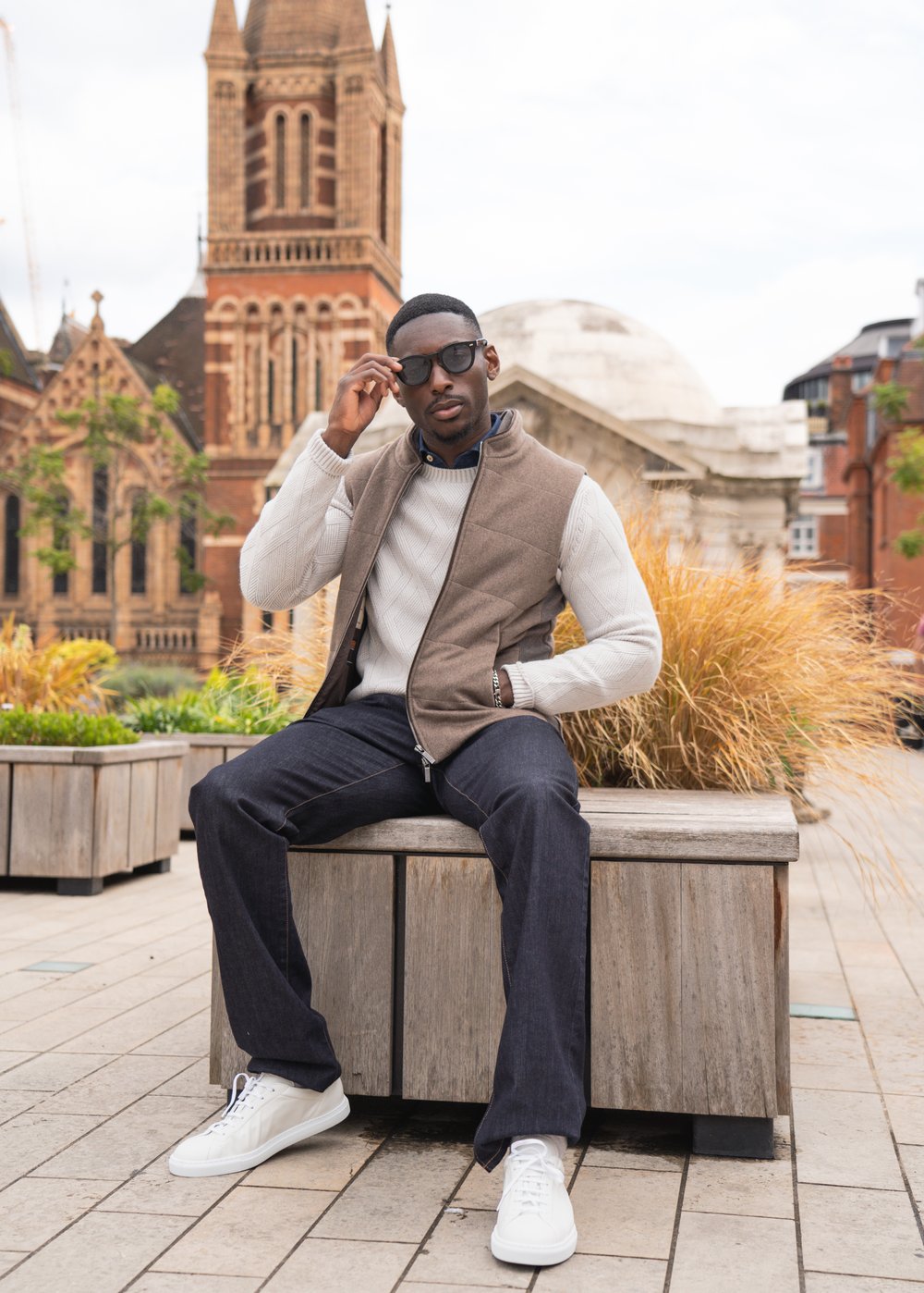 A man in sunglasses, a beige vest, white sweater, dark jeans, and white sneakers sits on a wooden bench outdoors. Behind him are tall grasses in planters, a brick church, and modern buildings under a cloudy sky. , available at Richard Gelding Mayfair