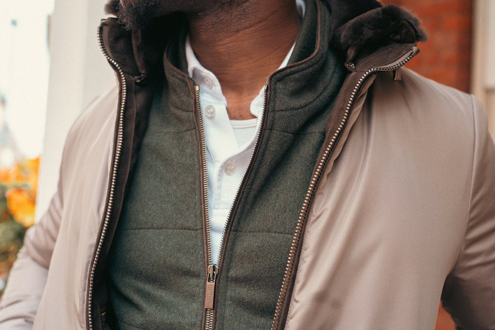A close-up of a person wearing a beige jacket with a fur-lined hood, a green zip-up vest, and a white button-up shirt. The jacket is unzipped, revealing the layered clothing underneath. The persons head is partially out of frame. , available at Richard Gelding Mayfair