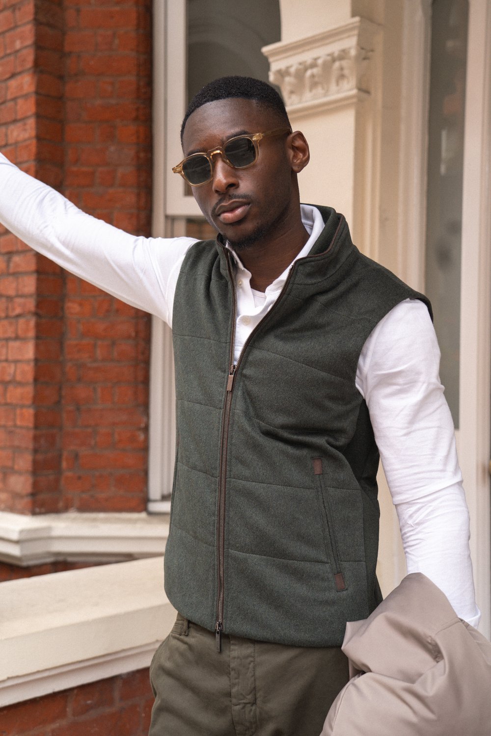 A man wearing sunglasses, a dark green zip-up vest over a white long-sleeve shirt, and olive pants stands outside a building with red brick and cream trim, holding a beige coat and resting one arm on a ledge. , available at Richard Gelding Mayfair