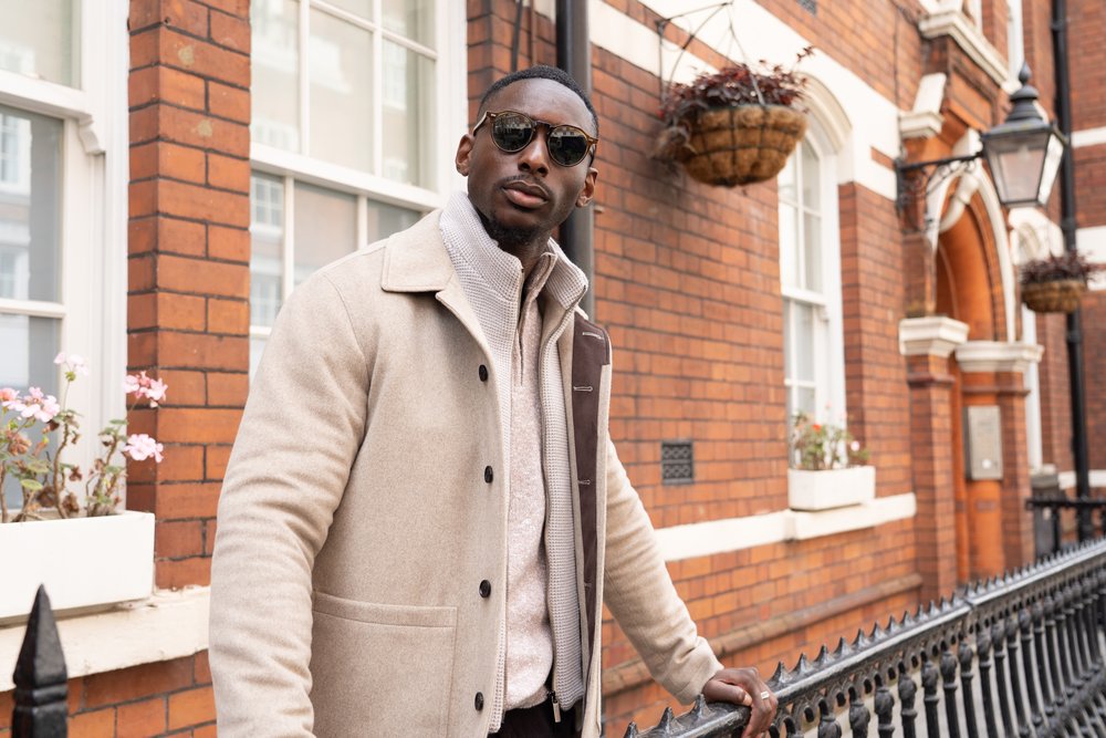 A man wearing sunglasses, a beige coat, and a light zip-up sweater stands confidently by a black iron fence in front of a red brick building with white-framed windows and hanging flower baskets. He looks directly at the camera. , available at Richard Gelding Mayfair