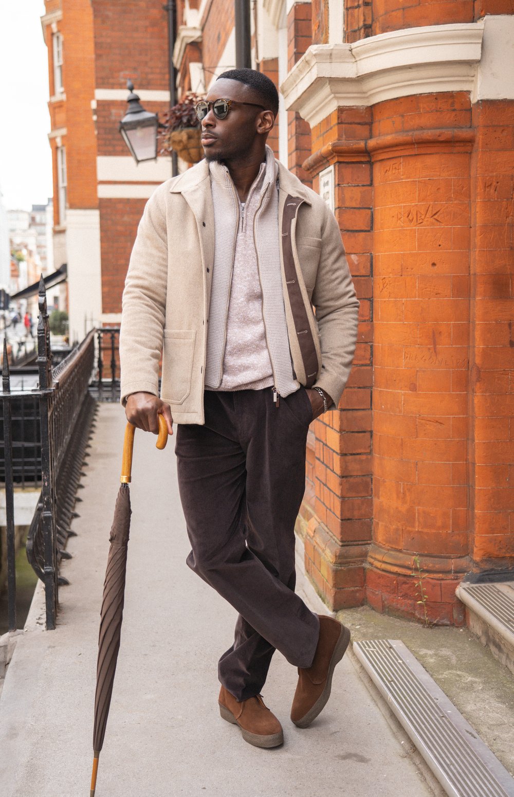 A man stands outdoors by a red brick building, wearing sunglasses, a beige jacket, light sweater, dark corduroy pants, and brown shoes. He holds a closed umbrella and looks to the side, exuding a relaxed, stylish vibe in an urban setting. , available at Richard Gelding Mayfair
