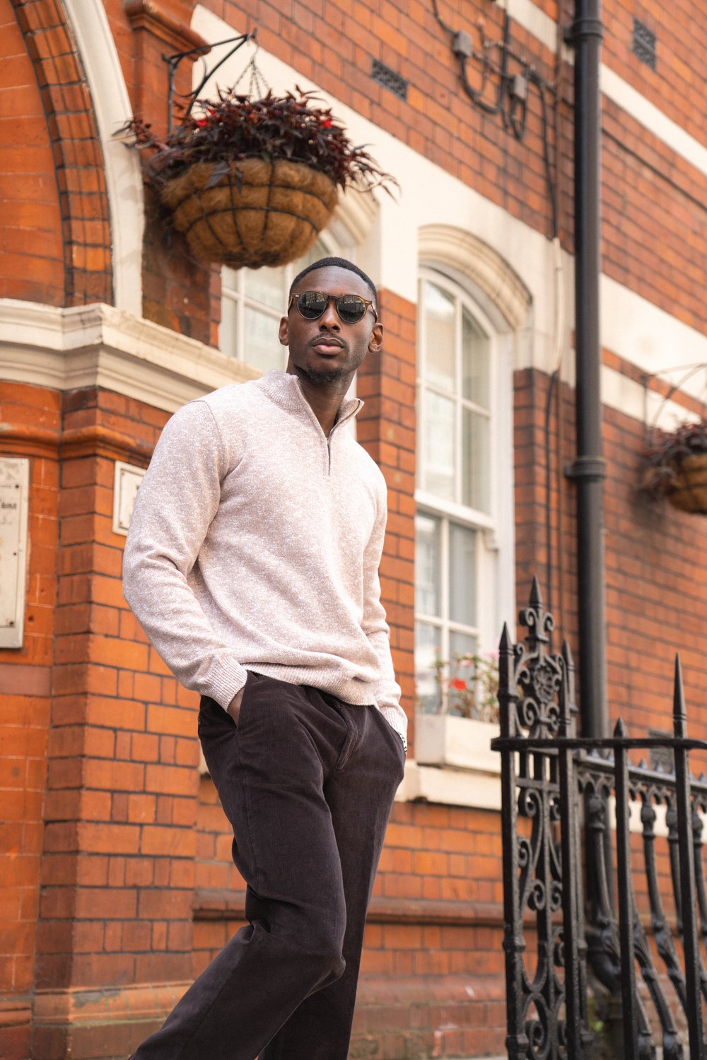 A man wearing sunglasses, a light beige pullover, and dark trousers stands confidently with hands in pockets in front of a red brick building with white trim, tall windows, and hanging flower baskets. Black iron railings are beside him on the sidewalk. , available at Richard Gelding Mayfair