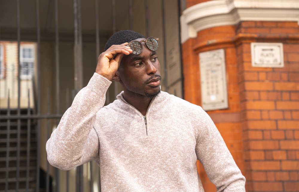A man in a light beige sweater lifts sunglasses from his eyes while looking to the side. He stands in front of a brick building with white trim and black metal gates, with plaques mounted on the wall behind him. , available at Richard Gelding Mayfair
