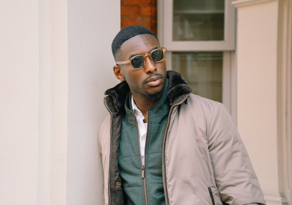 A stylish man with short hair and trimmed beard leans against a white wall. He wears round sunglasses, a light gray jacket with dark collar, and a green inner jacket with a white shirt underneath. He gazes to the side with a confident expression. , available at Richard Gelding Mayfair
