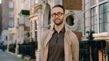 A man with short brown hair, a beard, and glasses stands on a city sidewalk. He wears a Gran Sasso Dark Brown Knitted Silk Zip Polo Shirt and a light tan jacket. Ornate brick and stone buildings with black railings rise behind him. , available at Richard Gelding Mayfair