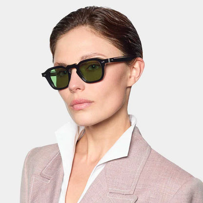 A woman with slicked-back brown hair wears TBD Eyewear's Ply Black | Green rectangular sunglasses and a light mauve blazer over a white shirt, facing slightly right against a plain background for a stylish, confident look. , available at Richard Gelding Mayfair