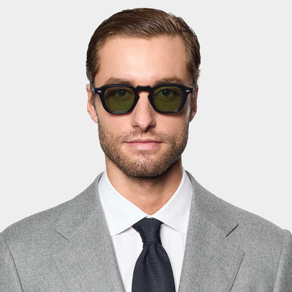 A man in a light gray suit, white shirt, and dark tie faces forward. He wears TBD Eyewear's Ply Black | Green handcrafted sunglasses with green lenses. He has short brown hair, a trimmed beard, and the background is plain and light-colored. , available at Richard Gelding Mayfair