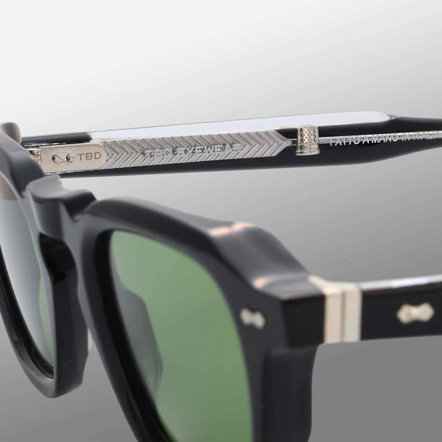 Close-up of Ply Black | Green by TBD Eyewear: black sunglasses with green lenses, handcrafted in Italy. Focus on the silver chevron-patterned arm with engraved text, visible hinge detail, metal accent, and emblem near the frame front. , available at Richard Gelding Mayfair