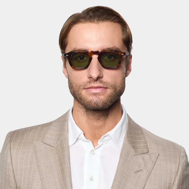 A fair-skinned man with light brown hair and a trimmed beard wears TBD Eyewear's Ply Brown Havana | Green sunglasses, featuring a handcrafted dark green frame, paired with a beige pinstripe suit and white shirt on a light gray background. , available at Richard Gelding Mayfair