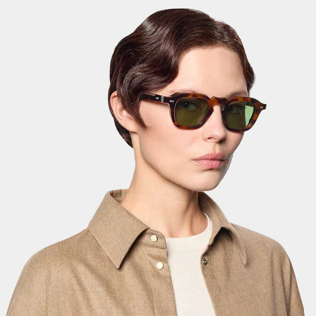 A person with short, wavy brown hair wears TBD Eyewear Ply Brown Havana | Green sunglasses with an acetate renew frame and a tan collared shirt over a white top, posing against a plain light gray background facing slightly right. , available at Richard Gelding Mayfair