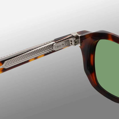 Close-up of TBD Eyewear Ply Brown Havana | Green sunglasses featuring green lenses in an acetate renew frame. The metal hinge has a chevron pattern and engraved logo. Handcrafted in Italy, shown on a smooth gradient gray background. , available at Richard Gelding Mayfair
