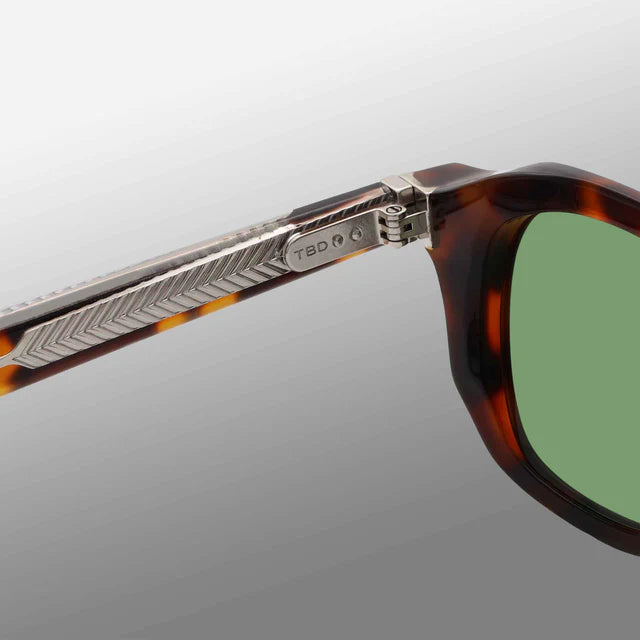 Close-up of TBD Eyewear Ply Brown Havana | Green sunglasses featuring green lenses in an acetate renew frame. The metal hinge has a chevron pattern and engraved logo. Handcrafted in Italy, shown on a smooth gradient gray background. , available at Richard Gelding Mayfair