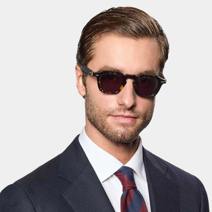 A man in a dark suit, white shirt, and red-and-blue striped tie wears TBD Eyewear Ply Black Havana | Grey handcrafted Italian rectangular sunglasses with dark lenses. He has short brown hair, a trimmed beard, and stands against a light gray background. , available at Richard Gelding Mayfair
