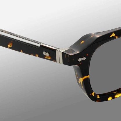 Close-up of Ply Black Havana | Grey eyeglasses by TBD Eyewear, handcrafted in Italy with a glossy black and yellow tortoiseshell pattern. The metal hinge joins the frame front and arm, set against a clean light gray gradient background. , available at Richard Gelding Mayfair