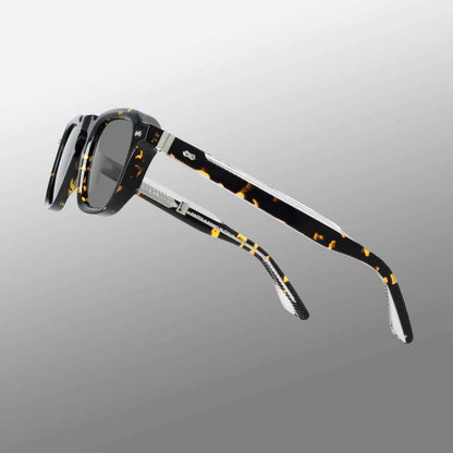 The Ply Black Havana | Grey sunglasses by TBD Eyewear are handcrafted in Italy with thick, premium acetate renew frames, dark grey-tinted lenses, and arms showcasing a mottled amber and black pattern with metallic accents. , available at Richard Gelding Mayfair