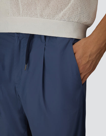 A person wears Herno Blue Lightweight Nylon Drawstring Casual Trousers with a pleated design, paired with a white textured knit sweater. Only the torso and upper legs are visible, set against a plain background. One hand rests in the pocket. , available at Richard Gelding Mayfair