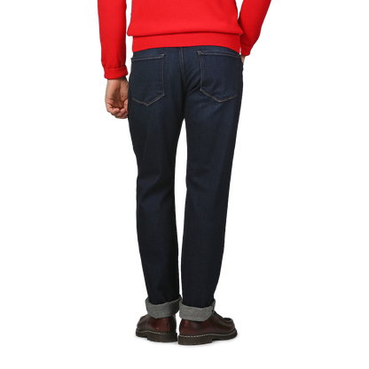 A person in a red sweater and Paige's Blue Faded Cotton Cramer Normandie Jeans stands with their back facing the camera, wearing brown shoes.