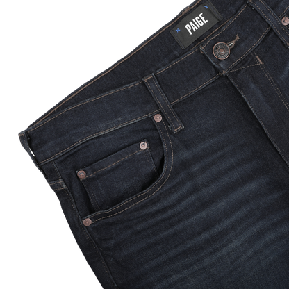 Close-up of dark blue denim Blue Faded Cotton Cramer Normandie Jeans featuring a visible "Paige" label on the waistband, showcasing front pocket and button details. These straight-leg jeans incorporate a hint of cotton stretch, providing comfort and style throughout the day.