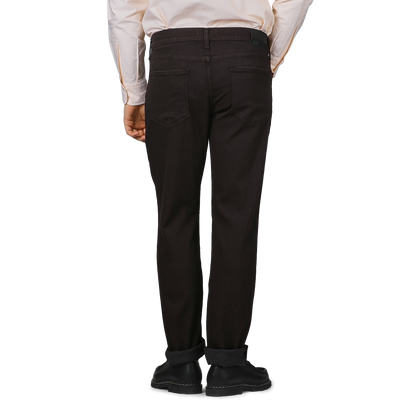 Rear view of a person wearing a striped shirt and Paige's Brown Cotton Transcend Normandie Jeans, hands by their sides against a gray background.
