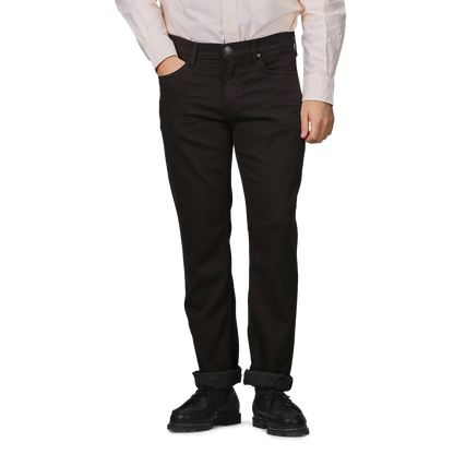 Wearing a white shirt and black shoes, a person stands against a plain background, embodying the elegance of Paige's Brown Cotton Transcend Normandie Jeans with folded cuffs.
