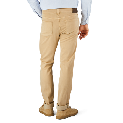 A person wearing beige pants with a comfortable fit and a light blue shirt is shown from the back. The Caramel Beige Cotton Transcend Federal Slim Jeans, possibly by Paige, have two back pockets. They are also wearing brown shoes.