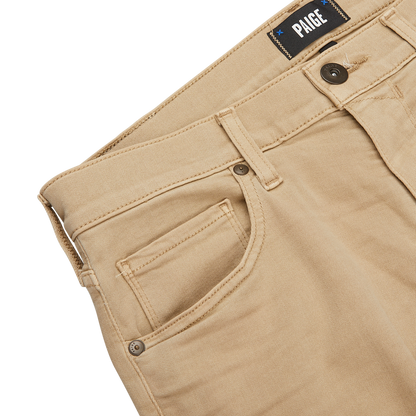 Close-up of beige pants with a visible label reading "PAIGE" above the waistband, showcasing belt loops, front pocket, and button closure on a light surface. These Caramel Beige Cotton Transcend Federal Slim Jeans are crafted from Transcend fabric for a comfortable fit.