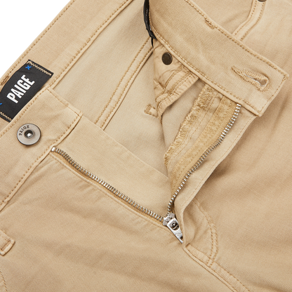 Close-up of Caramel Beige Cotton Transcend Federal Slim Jeans with a partially open zipper and button. The brand label "Paige" is visible above the pocket, showcasing the comfortable fit and luxurious Transcend fabric.