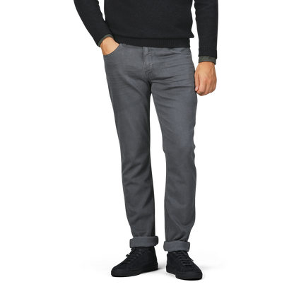 A person wearing a black long-sleeve shirt, black shoes, and Paige Charcoal Cotton Transcend Federal Slim Jeans in onyx grey stands with hands in front pockets. The jeans, made from comfortable Transcend fabric, are rolled up at the cuffs. The head and upper body are not visible.