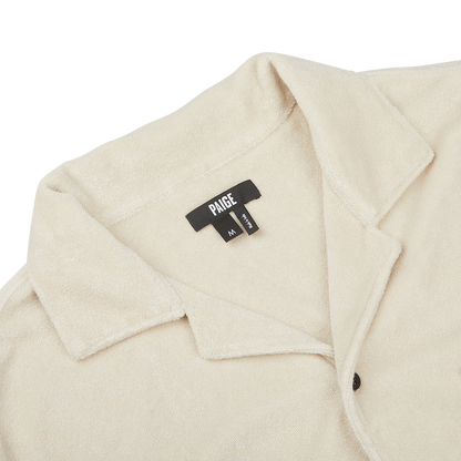 Close-up of a Cream Beige Cotton Towelling Polo Shirt with a front button opening, collared design, and a black fabric tag displaying the brand "Paige" and size "M".
