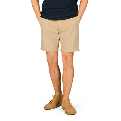 A person standing with their hands behind their back, wearing a navy blue t-shirt, Paige Khaki Beige Cotton Blend Phillips shorts, and light brown shoes.