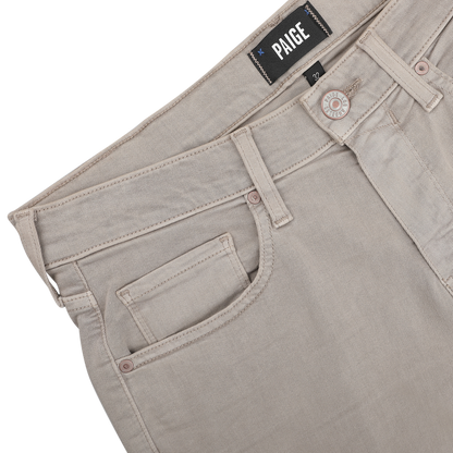The Moroccan Clay Cotton Transcend Federal Slim Jeans, crafted by Paige, showcase a unique Moroccan clay color and feature light beige denim. The jeans include a front pocket with a visible button and the "Paige" label elegantly displayed on the waistband, enhancing their versatile style.