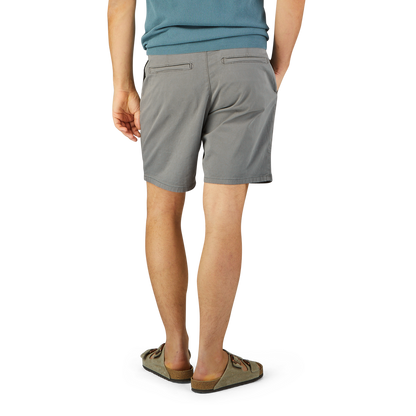 A man wearing Paige's slate grey cotton blend Phillips shorts and beige slip-on shoes stands with his back to the camera against a blue and yellow background.
