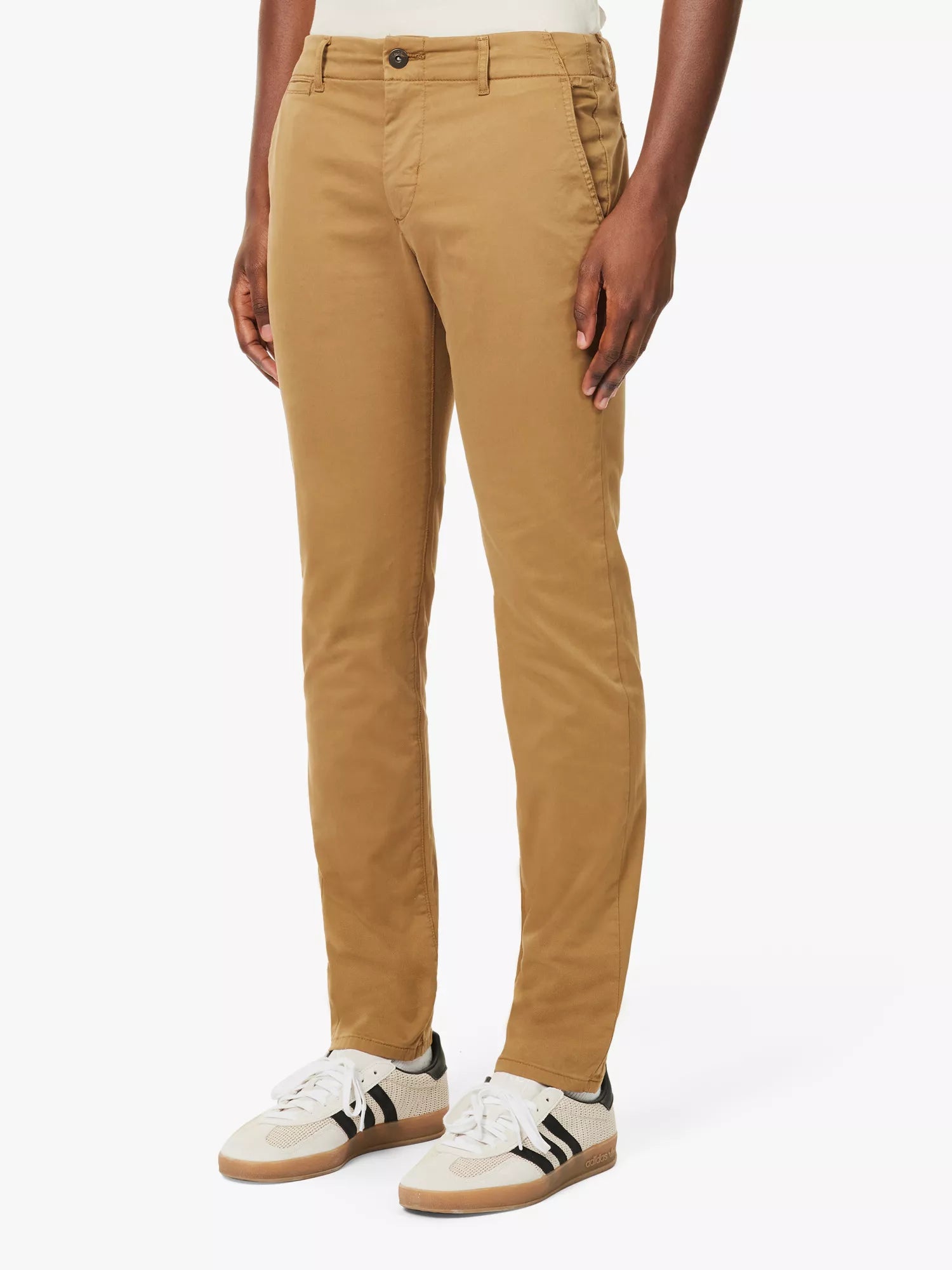 A person stands against a white background, wearing Paige’s Tobacco Brown Danford Cotton Chino with a cream shirt tucked in and white sneakers with black accents. Only the lower half of their body is visible, hands relaxed at their sides. , available at Richard Gelding Mayfair