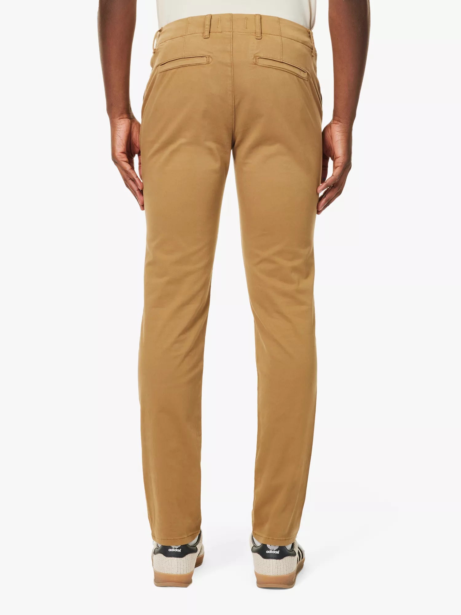A person faces away from the camera, wearing Paige's Tobacco Brown Danford Cotton Chino with slim fit, buttoned back pockets, and belt loops. They pair the chinos with black and white sneakers against a plain white background. , available at Richard Gelding Mayfair