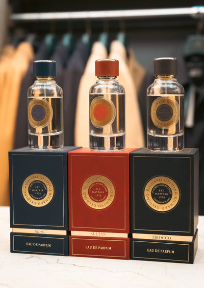 Three glass perfume bottles with colored caps rest on boxes, including Richard Gelding Sirocco EdP 100ml. Blurred coats in the background highlight Sirocco's long-lasting, oriental fragrance by Richard Gelding. , available at Richard Gelding Mayfair