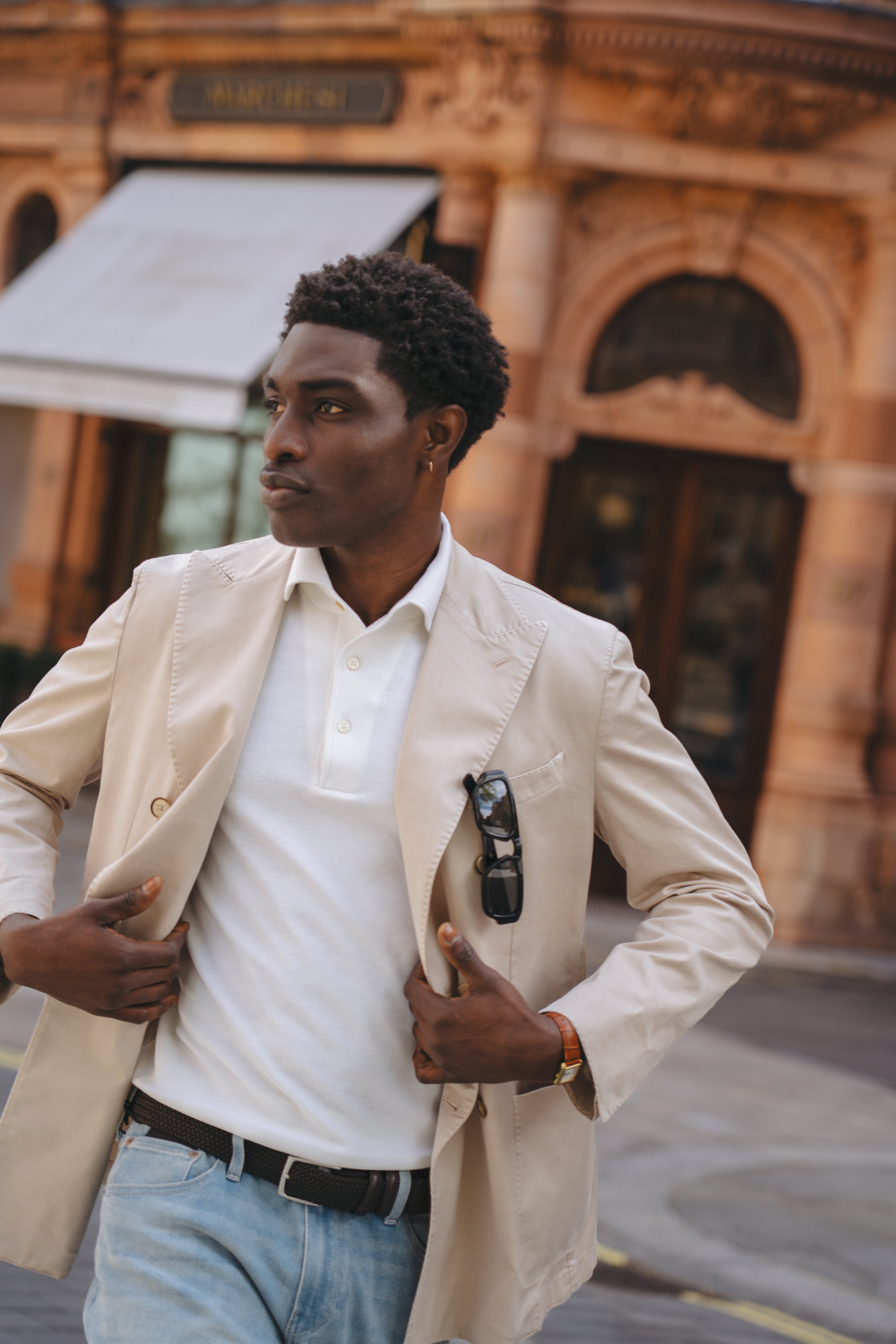 A stylish man outdoors in front of arched windows adjusts his Fedeli Beige Cotton Double Breasted Blazer, worn over a white polo and light blue jeans, with sunglasses hooked onto the lapel amid warm-toned architecture. , available at Richard Gelding Mayfair