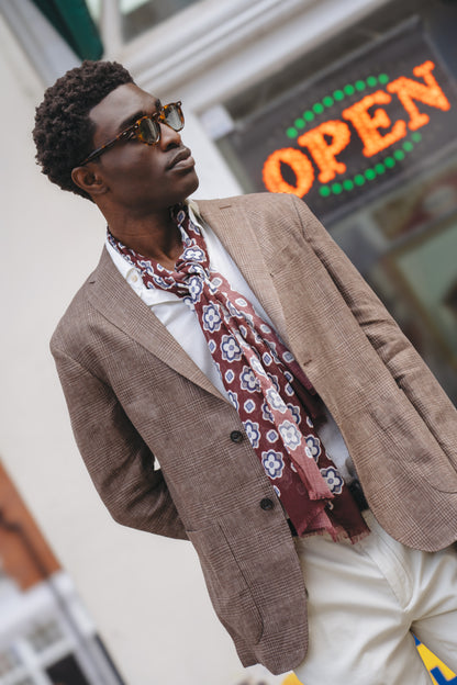 A stylish man in sunglasses wears a Boglioli Brown Prince Of Wales Check Linen Jacket, white shirt, cream pants, and maroon scarf. He stands outside with hands in pockets, gazing left as a neon "OPEN" sign glows behind him. , available at Richard Gelding Mayfair