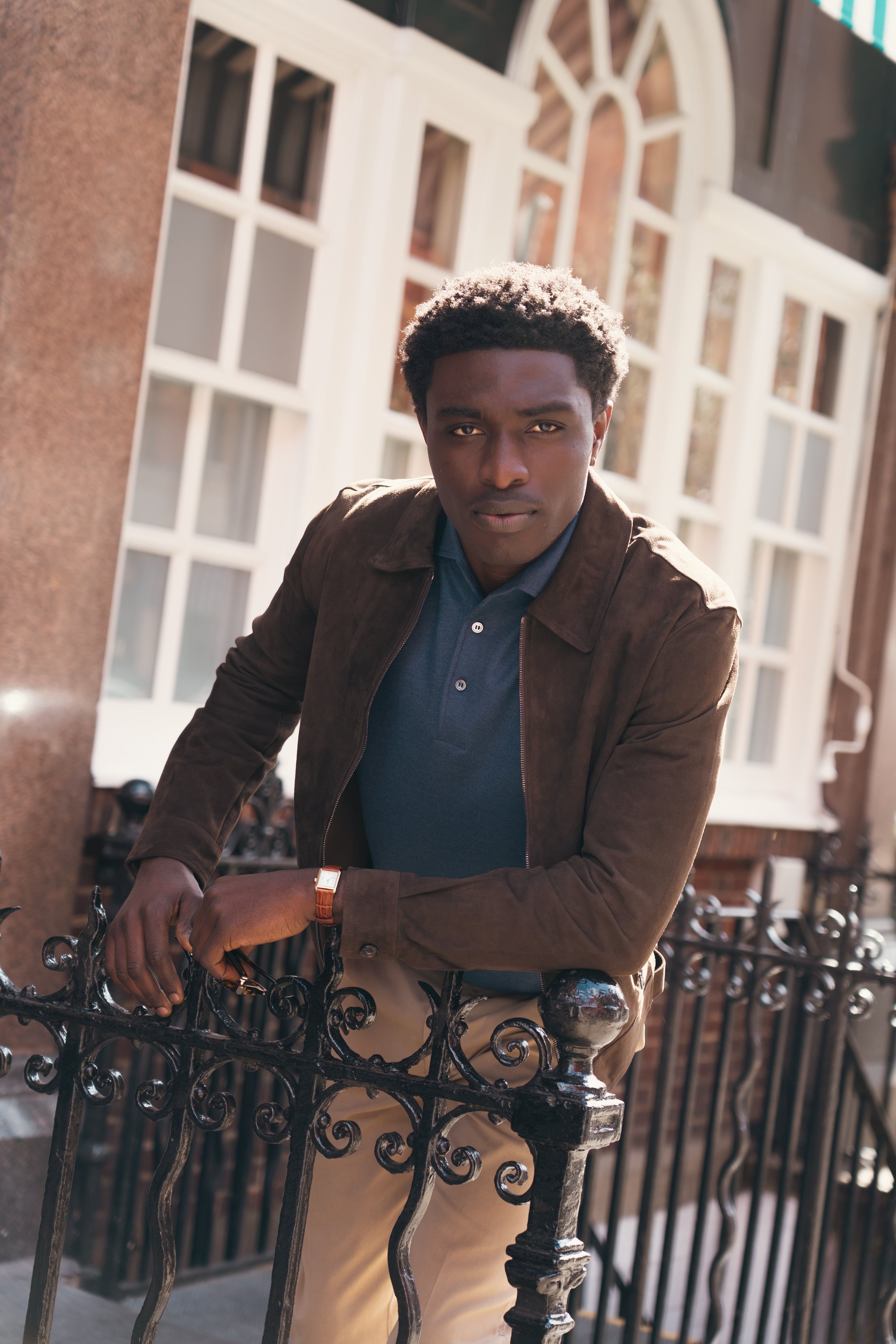 A man with short curly hair leans on a black iron fence outside, wearing the Manto Chocolate Brown Strozzi Zip Suede Blouson over a navy polo and beige pants. Sunlight highlights large white-framed windows and a stone building behind him. , available at Richard Gelding Mayfair